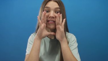 Genç, beyaz bir kız, parlak mavi bir stüdyoda bağırırken iki elini de ağzına dayar..