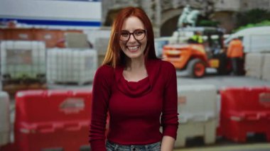 İnşaat alanında gülen kızıl saçlı, kırmızı kazaklı ve gözlüklü bir kadın gün boyunca inşaat bariyerleri ve makineleri karşısında gülümsüyor..