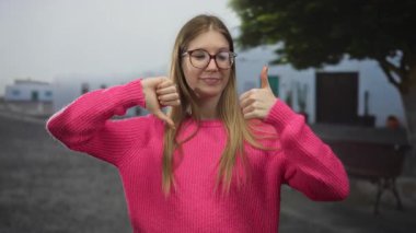 Pembe kazak giyen gülümseyen sarışın kadın, güneşli caddede baş parmağını kaldırır ve baş parmağını kaldırır; kararsızlık.
