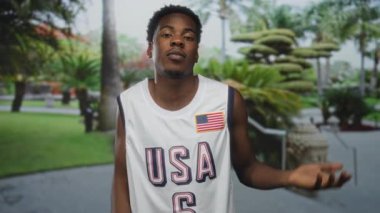 Adam, yemyeşil parkta beyaz Amerikan basketbol forması giyip, Amerikan bayrağıyla süslenmiş bir şekilde elini kaldırıyor..