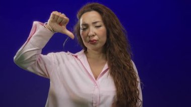 Mavi stüdyoda pembe gömlek ve bilezikler giyen, baş parmağıyla ve eliyle direkt bakışlarla işaret eden bir kadın..