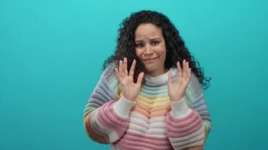 Renkli süveter giyen genç İspanyol kadın dışlanmış mavi arka plana karşı anlamlı duygularla işaret ediyor.