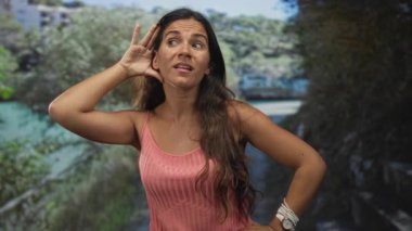 Kulağını elleriyle kavrayan, çıplak kulağı görünen, su ve binanın yanındaki orman yolunda pembe kolsuz bluz giyen bir kadın..