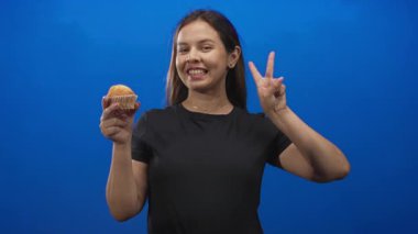 Mavi stüdyoda barış işareti yaparken gülümseyen ve kek tutan genç kadın; neşeli atıştırmalık kutlaması.