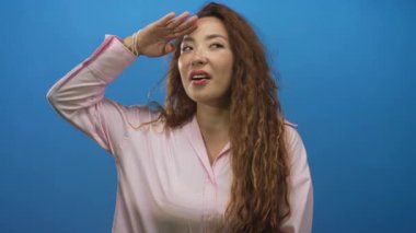 Mavi stüdyoda eli yüzü üzerinde, uzun kıvırcık saçlı ve pembe bluzlu kadın koruyucu gözleri görünür; beklenti.