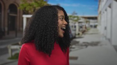 Kıvırcık saçlı, kırmızı tişörtlü, güpegündüz gülümseyen dişleri olan, neşeli iyimser bir kadın..