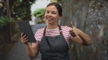 Kot önlüklü gülümseyen kadın tableti tutuyor ve taş duvarlı dar bir sokakta baş parmak kaldırıyor. Küçük işletme onayı..