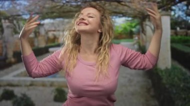 Orman pergola geçidinde kollar açıkken elleri ile kalp oluşturan ve pembe bir bluz içinde gülümseyen kadın; neşe kurtuluşu.