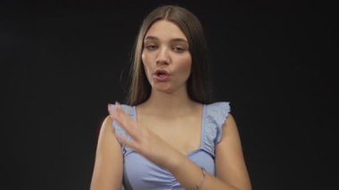 Açık mavi tanktop giyen bir kız siyah stüdyoda dudaklarını büzüp göz teması kurarken çıplak kollarını çaprazlıyor..