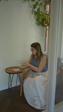 Kadın, rahat bir kafeteryada, içinde küçük bir masa ve sallanan bir bitkinin bulunduğu, rahat bir kafeteryada kahveden zevk alıyor. Huzurlu bir ortam yaratıyor..