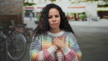 İşlek bir sokakta gökkuşağı süveteri giyip ellerini göğsünün önünde tutan genç İspanyol kadın arka planda bisiklet ve yayalar bulanıklaşırken,.