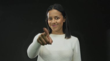 Gülümseyen bir kadın, siyah bir arkaplana işaret ederken, bir stüdyo ortamında ifade ve duygulara odaklanmış bir şekilde bağlanma ve güven iletiyor..