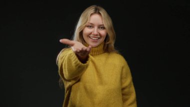 Siyah arka planda sarı bir kazakla gülümseyen bir kadın elini uzatıyor genç sarışın bir kadın arkadaşlığını ve sıcaklığını gösteriyor..