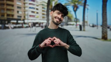 Genç adam güneşli bir şehir sokağında yeşil bir kazak giymiş, kameraya gülümseyen, sıcak ve misafirperver bir atmosfer yaratan bir kalp hareketi yapıyor..