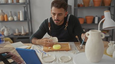 Kil kasesini çıplak elle şekillendiren adam, sanat atölyesinde çömlek raflarıyla ahşap masa üzerinde, yaratıcılığa odaklanıyor..