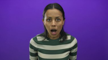 İspanyol kadın, yeşil çizgili gömleği olan, izole edilmiş mor bir arka plan duvarında dururken şaşkınlığını dile getiriyor..