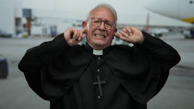 Kıdemli rahip, yoğun havaalanı arka planında hayal kırıklığı içinde yüzükoyun yatıyor, elleri kulaklarını tıkıyor, büyük haç ve gündüz sahneleriyle dini giysiler giyiyor..