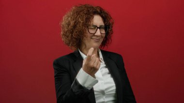 Orta yaşlı, takım elbiseli İspanyol bir kadın, canlı kırmızı bir duvara el hareketi yaparak izole edilmiş bir stüdyoda güven ve iletişimi harmanlıyor..