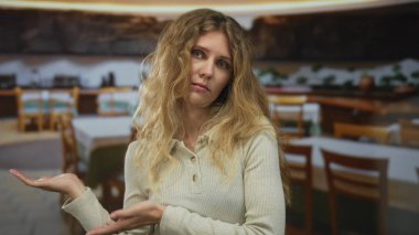 Kadın, restoranda omuz silkerek el kaldırıyor. Sarışın, genç yüzü dağınık ve belirsiz bir ifade takınıyor..
