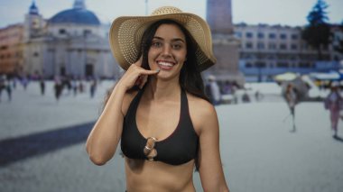 Hasır şapka ve bikinili kadın, Vatikan meydanında güneş ışığı altında çıplak omuzlu çağrı jesti yapıyor; bağlantı neşesi.