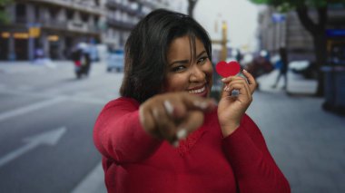 Kırmızılı Latin kadın kalp şeklinde bir kağıt tutuyor, hareketli bir sokak şehrinin arka planında güvenle gülümsüyor, açık havada sevgi ve pozitifliği somutlaştırıyor..