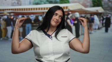 Elleri açık şehir pazarında poz veren kadın, insanların alışveriş ve hareketli market tezgahlarının arka planına karışıyor..