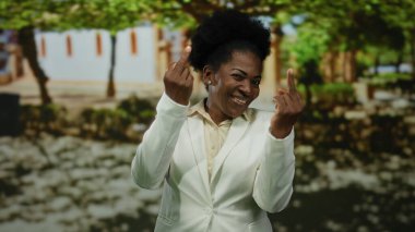 Parkta gülümseyen bir kadın arka planda ağaçlar ve gölgelerle el hareketleriyle dışarıdaki kaygısız sahneyi vurguluyor..