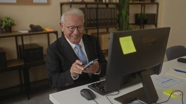 Modern ofis içinde akıllı telefon ve bilgisayar kullanan kıdemli bir adam. İşyerinde raflar ve temiz bir masa ile odaklanmış ve meşgul görünüyor..