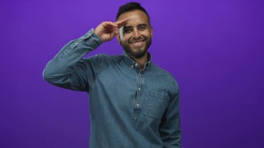 Kot gömlekli genç İspanyol adam mor bir arka plana karşı stüdyoda el pençe divan gülüyor ve selam veriyor..