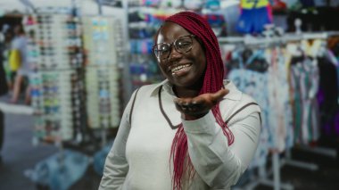 Saçları örgülü bir kadın hareketli bir pazar caddesinde kameraya öpücük yolluyor, renkli malların arasında güvenle gülümsüyor açık hava alışveriş atmosferini güzelleştiriyor..