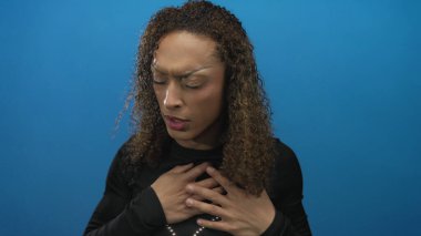 İspanyol transseksüel kadın göğsünü kapalı gözlerle tutuyor ve mavi stüdyoda kaşlarını çatıyor; acı kalp krizi.