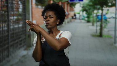 Önlüklü bir kadın, şehir sokaklarında hareketli ve kendinden emin bir şekilde, hayat dolu arka planlarla dolu bir şehir ortamında el tabancası jesti yapıyor..