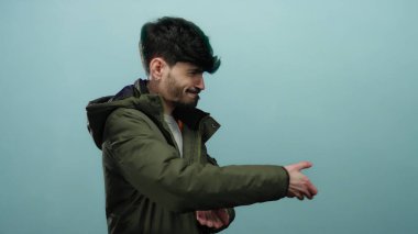 Mavi bir arka plana hayali bir ip çeken genç bir adam, yeşil bir ceket giyiyor ve neşeli görünüyor, yüzünde odaklanmış bir ifadeyle çevreden soyutlanmış..