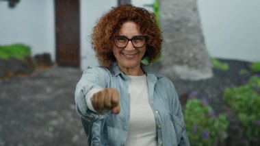 Kıvırcık saçlı İspanyol kadın şehir caddesinde kameraya yumruğunu sergiliyor. Açık hava ortamında güç ve pozitifliği somutlaştırıyor..