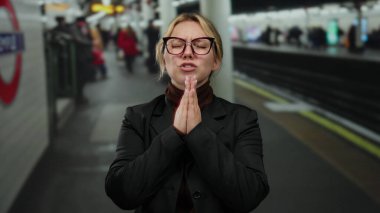 Yoğun tren istasyonunda gözlükleriyle dua eden bir kadın, konsantrasyonunu ve umudunu banliyö istasyonunun ortasındaki kapalı bir tren istasyonunda aktarıyor..