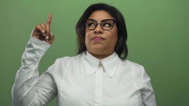 Genç Latin bir kadın yeşil bir duvarın önünde tiksindirici bir ifadeyle yukarıya doğru işaret ediyor..