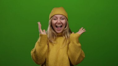 Parlak yeşil bir altyapının önünde neşe saçan, rahat sarı bir süveter giyen, sarı saçlı ve neşeli bir gülümsemesi olan bir kadın..