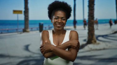 Afrika kökenli Amerikalı bir kadın, arka planda palmiye ağaçları ve okyanusla güneşli bir sahil yürüyüşünde sıcaklığını ve kendini sevgini ifade ediyor..