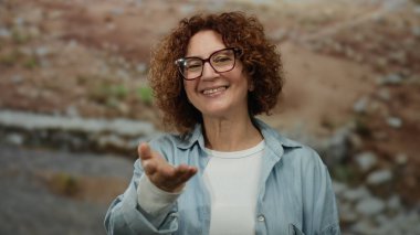 İspanyol orta yaşlı bir kadın şehir sokaklarında dostça bir gülümsemeyle elini uzatıyor, gözlük takıyor ve günlük giysiler giyiyor, şehir manzarası ortamında etkileşime girmeye davet ediyor..