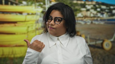 Beyaz tişörtlü ve gözlüklü, iri yarı, Latin bir kadın sahil kenarındaki arka plana doğru başparmağıyla güvenle duruyor..