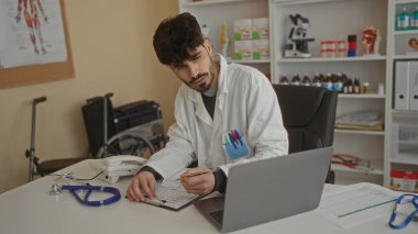 Doktor, klinikte steteskop ve dizüstü bilgisayarla tıbbi ekipman ve evraklarla çevrili halde profesyonel bir sağlık ortamını gösteren notlar yazıyor..