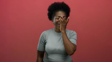 Yüzü koyun gülen bir kadın, izole edilmiş kırmızı arka plana karşı duruyor, neşe ve şaşkınlık ifade ediyor, afro saç stili, gri gömlek, genç Afro-Amerikan.