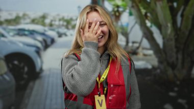 Şehirde bir kadın sokakta gönüllü olarak gülerken bulutlu gökyüzünün altında kırmızı yelek ve sarı rozet takıyor..