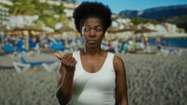 Sahilde arka planda deniz ve şemsiyelerle baş parmağını gösteren bir kadın, açık gökyüzü altında gevşek deniz havasını yakalıyor..