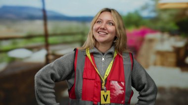Kırmızı yelek giyen ve gönüllü rozet takan bir kadın şehirdeki hareketli bir açık hava restoranının terasında dikiliyor. Etrafı canlı çiçekler ve şehir manzarasıyla çevrili..