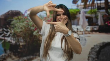 Güneşli bir sahilde gülümseyen bir kadın elleri parlak çiçekler ve açık mavi gökyüzü ile çevrili bir çerçeve hareketi yapıyor.