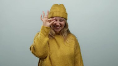 Sarı kazaklı, şapkalı, gülümseyen bir kadın beyaz arka planda izole edilmiş el hareketi yaparak neşeli, olumlu bir ifade ve kış modasında samimiyet gösteriyor..