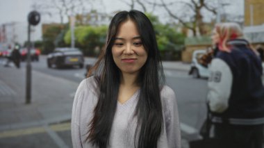 Uzun saçlı bir kadın sokakta yüz ifadeleri yapıyor. Bulanık arabaları ve yeşillikleri var. Açık hava ortamında çeşitli tepkiler gösteriyor..