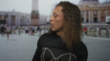 Kadın başını çevirir ve bina yakınında ağzını açar kıvırcık saçlar yüzünü çerçeveler ve çivili bir üst giyer;.