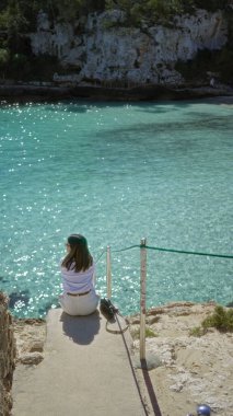 Mallorca 'daki Cala Lombars' da turkuaz su kenarında oturan kadın, güneşli ve huzurlu bir atmosfer olan Akdeniz manzarasının tadını çıkarıyor..